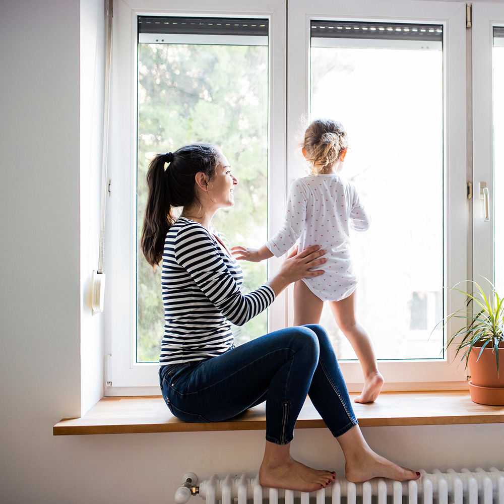 Beautiful young mother sitting on window sill with her cute little daughter looking out of window